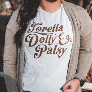 May include: A light-colored t-shirt with the words "Loretta Dolly & Patsy" in brown script. The shirt is worn with a beige cardigan and blue jeans. A watch is visible on the wrist.