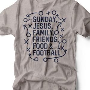 Puede incluir: Una camiseta gris claro con un diseño gráfico azul marino. El diseño incluye las palabras "SUNDAY, JESUS, FAMILY, FRIENDS, FOOD & FOOTBALL" con diagramas de jugadas de fútbol. La camiseta es de un material suave.