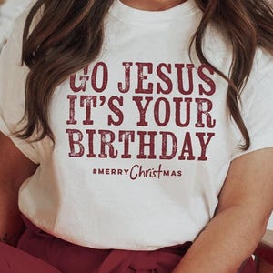 Puede incluir: Una camiseta blanca con el texto "GO JESUS IT'S YOUR BIRTHDAY" en letras burdeos. La frase "#MERRY Christmas" está impresa debajo. La camiseta se combina con pantalones burdeos.