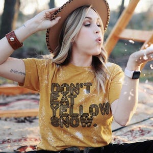 May include: Mustard yellow t-shirt with the text "DON'T EAT YELLOW SNOW." The shirt has a bleached design. The person is wearing a brown hat with a leopard print brim and a brown leather bracelet.