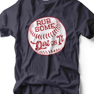 May include: Navy blue t-shirt featuring a baseball graphic with the text "Rub Some Dirt On It" in red. The baseball design is white with red stitching. The shirt is a casual wear item.