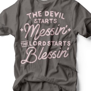 Puede incluir: Camiseta gris oscuro con el texto "THE DEVIL STARTS Messin' + THE LORD STARTS Blessin'" en escritura rosa claro. La camiseta tiene cuello redondo y mangas cortas. La marca "RUBY'S RUBBISH" está en la parte inferior.