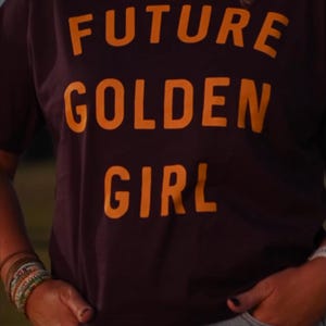 May include: A dark purple t-shirt with the words "FUTURE GOLDEN GIRL" in bright orange block letters. The person is wearing blue jeans and has curly blonde hair. The person's hands are in their pockets.
