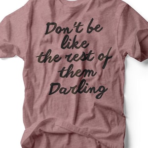 May include: A dusty rose t-shirt with the text "Don't be like the rest of them Darling" in black script. The shirt is made of a soft material and has a crew neck. The brand name "RUBY'S RUBBISH" is printed at the bottom.