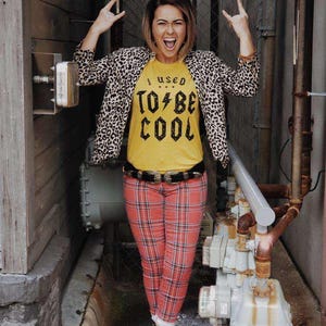 Può includere: Una persona indossa una maglietta gialla con la scritta "I USED TO BE COOL", una giacca con stampa leopardata e pantaloni a quadri rossi. Indossa una cintura nera e stivali neri. Sta facendo un gesto rock and roll con la mano.