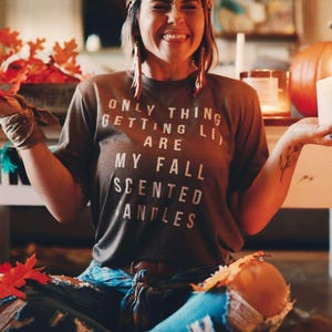 Op de afbeelding: Een vrouw draagt een bruin t-shirt met de tekst "ONLY THING GETTING LIT ARE MY FALL SCENTED CANDLES". Ze draagt een luipaardprint hoofdband en een gescheurde blauwe jeans. De afbeelding heeft een herfstthema.