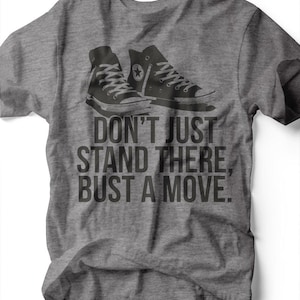 Op de afbeelding: Grijze t-shirt met een afbeelding van twee zwarte high-top sneakers. De tekst "DON'T JUST STAND THERE, BUST A MOVE" staat eronder in donkergrijs. Het shirt is gemaakt van een zacht materiaal.