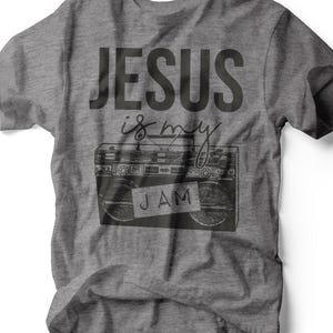 Puede incluir: Camiseta gris con el texto "JESUS is my JAM" en negro. El diseño incluye un gráfico de un radiocasete. La camiseta está hecha de un material suave y es adecuada para uso casual.