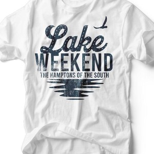 Puede incluir: Camiseta blanca con las palabras "Lake Weekend" en una fuente azul oscuro texturizada. Debajo, el texto "The Hamptons of the South" está impreso. Un gráfico estilizado de un pájaro está encima de la palabra "Weekend", y un gráfico de reflejo de lago debajo.