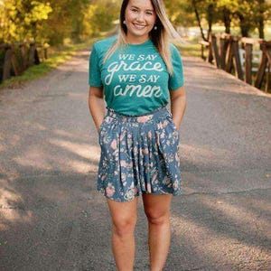 Puede incluir: Una mujer con una camiseta verde azulado con la frase "We say grace, we say amen" en escritura blanca. También lleva una falda azul floral y botines marrones. El fondo es una escena borrosa al aire libre.