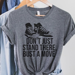 Op de afbeelding: Een heather grijze t-shirt met een zwarte afbeelding van hoge sneakers en de tekst "DON'T JUST STAND THERE, BUST A MOVE." Het shirt hangt aan een houten hanger met andere kleding.