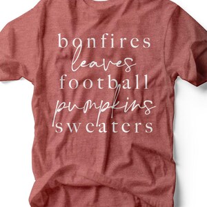 May include: A red-orange t-shirt with the words "bonfires, leaves, football, pumpkins, sweaters" printed in white. The shirt has a crew neck and short sleeves. The text is in a mix of fonts.