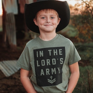 May include: A young person wearing a black cowboy hat and a green t-shirt with the text "IN THE LORD'S ARMY" in a black rectangle. The person is also wearing blue jeans.