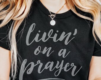 Peut inclure: T-shirt noir avec l'inscription "Livin' on a Prayer" en gris clair et en écriture cursive. Le col est rond. Un collier doré avec un pendentif est visible.