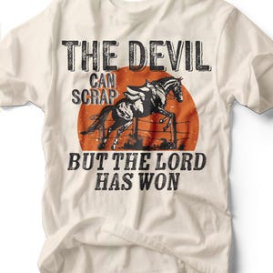 May include: Off-white t-shirt featuring a black and white illustration of a horse jumping over a fence, set against an orange sun. The text "THE DEVIL CAN SCRAP BUT THE LORD HAS WON" is printed above and below the image.