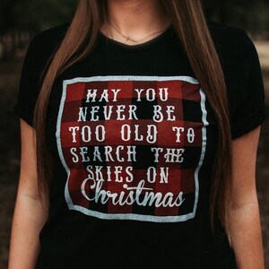 May include: Black t-shirt with a red and black plaid design and the text "MAY YOU NEVER BE TOO OLD TO SEARCH THE SKIES ON Christmas" in white. The shirt is a crew neck and has short sleeves.