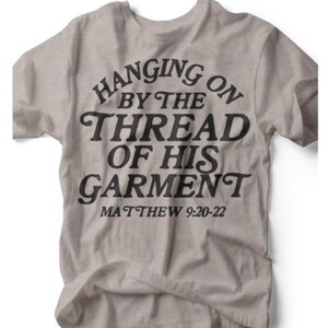 Puede incluir: Camiseta gris claro con el texto negro "HANGING ON BY THE THREAD OF HIS GARMENT" y "MATTHEW 9:20-22". La camiseta tiene cuello redondo y está hecha de un material suave.