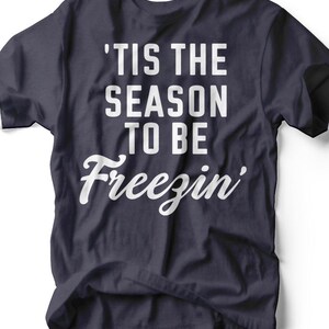 以下が含まれることがあります： 紺色のTシャツに、白い文字で「'Tis the Season to be Freezin'」と書かれています。このフレーズは、ブロック体と筆記体のフォントを組み合わせています。このシャツはカジュアルウェアです。