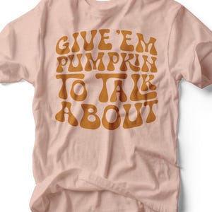 May include: A light pink t-shirt with the text "GIVE 'EM PUMPKIN TO TALK ABOUT" in orange lettering. The shirt is made of a soft material and is likely for casual wear. The design has a retro, groovy style.