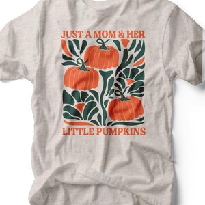 May include: A light gray t-shirt featuring a graphic design with three orange pumpkins and green foliage. The text "JUST A MOM & HER LITTLE PUMPKINS" is printed in orange. The shirt is made of a soft, comfortable material.