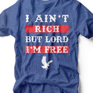 Può includere: T-shirt blu con la scritta bianca "I AIN'T RICH BUT LORD I'M FREE", con barre orizzontali rosse dietro le parole. Sotto c'è una grafica di un'aquila bianca. La maglietta è in materiale morbido.