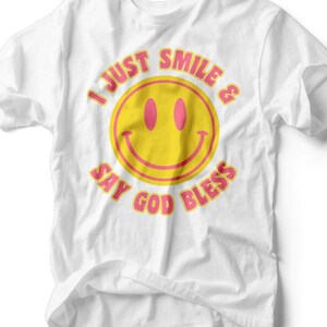 Puede incluir: Camiseta blanca con una gran carita sonriente amarilla y el texto "I JUST SMILE & SAY GOD BLESS" en rosa. La camiseta tiene cuello redondo y mangas cortas. El nombre de la marca "RUBY'S RUBBISH" está impreso en la parte inferior.