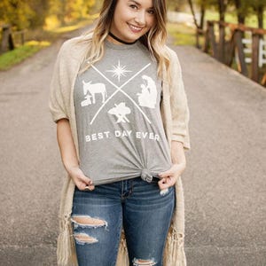 Puede incluir: Una mujer con una camiseta gris con un gráfico blanco y el texto "BEST DAY EVER". También lleva vaqueros azules rotos y una rebeca beige. El fondo es una escena borrosa al aire libre.