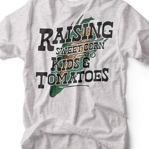 May include: A light gray t-shirt with the words "RAISING SWEET CORN KIDS & TOMATOES" in black text. The design includes a green and yellow corn graphic. The shirt is a casual wear item.