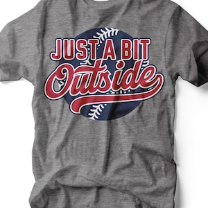 May include: Gray t-shirt with a baseball-themed graphic. The design features the words "JUST A BIT Outside" in red and white lettering over a blue baseball graphic. The shirt is made of a soft material.