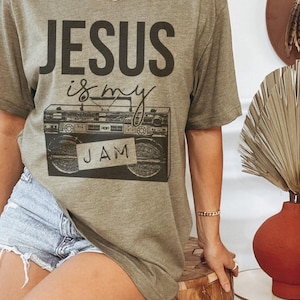 Puede incluir: Camiseta verde oliva con la frase "JESUS is my JAM" en negro. El diseño presenta un radiocasete vintage con una etiqueta "JAM" escrita a mano. Una prenda casual para el día a día.