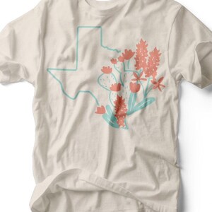 Könnte beinhalten: Cremefarbenes T-Shirt mit türkisfarbener Umrisslinie des Bundesstaates Texas. Das Design umfasst korallenfarbene Blumen und einen Kaktus mit türkisfarbenen Akzenten. Das Shirt hat einen Rundhalsausschnitt und kurze Ärmel. Der Text "RUBY'S RUBBISH" ist unten aufgedruckt.