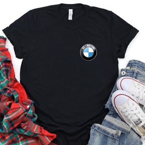 May include: Black t-shirt with a blue and white BMW logo on the front pocket. The shirt is laid out on a white background with a red and green plaid shirt, blue jeans, and white sneakers.