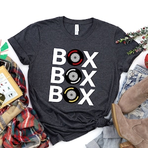 May include: A dark gray t-shirt with the word "BOX" repeated three times in white letters. Each "BOX" has a stylized black and white tire with red and yellow accents.