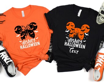 May include: Two t-shirts, one orange and one black, with a Halloween design. The design includes a silhouette of Mickey Mouse ears with a bow, bats, and a castle, and the words "Disney Halloween".