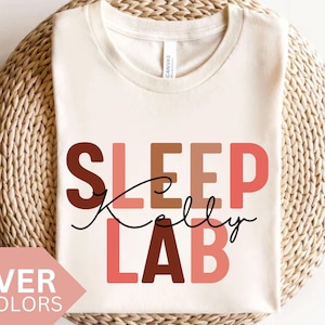 Può includere: Una maglietta bianca con il testo "SLEEP Kelly LAB" in lettere maiuscole colorate. Le lettere sono in tonalità di rosa, arancione e marrone.