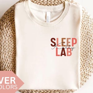 Può includere: Una maglietta bianca con il testo "SLEEP REALLY LAB" in un design impilato e colorato. Il testo è in tonalità di marrone, rosa e rosso. La maglietta è su una superficie di paglia intrecciata.