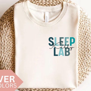 Może przedstawiać: Biały t-shirt z napisem "SLEEP Kelly LAB" w niebieskich i zielonych literach. Koszulka leży na plecionym podłożu.