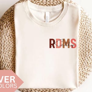 Könnte beinhalten: Ein weißes T-Shirt mit dem Text "RDMS" in Braun, Rosa und Orange. Der Text ist mit einer Herzform im "D" stilisiert. Der Text "Kelly" ist in schwarzer Kursivschrift unter dem "RDMS" geschrieben.