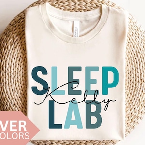 Può includere: Una maglietta bianca con il testo "SLEEP Kelly LAB" in blu e turchese. Il testo è in uno stile di font a blocchi.