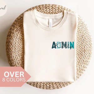 Könnte beinhalten: Ein weißes T-Shirt mit dem Text "ADMIN" in Blau, Grün und Türkis. Der Text ist mit einem Farbverlaufseffekt und einer schwarzen Umrandung gestaltet. Das Shirt liegt auf einer geflochtenen Weidenoberfläche.