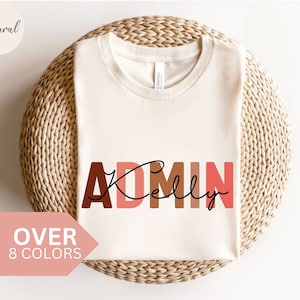 May include: A white t-shirt with the text "ADMIN Kelly" in brown, orange, and pink letters. The text is in a blocky font and the name "Kelly" is written in cursive.
