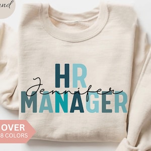 Peut inclure: Sweat-shirt couleur sable avec l'inscription "HR Jennifer MANAGER" en bleu sarcelle et bleu foncé. Une bannière rose indique "OVER 8 COLORS". Design épuré et moderne.