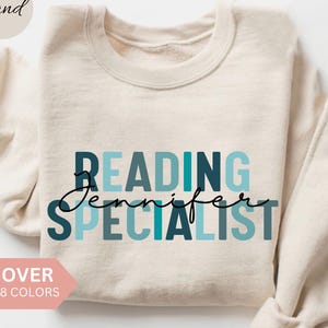 May include: Sand-colored sweatshirt with the words "READING SPECIALIST Jennifer" in teal and blue lettering. The sweatshirt has a crew neck and long sleeves. The text "OVER 8 COLORS" is in a pink box.