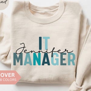 Puede incluir: Sudadera color arena con el texto "Jennifer IT MANAGER" en letras turquesas y azules. La palabra "Jennifer" está en cursiva. Una pancarta rosa dice "OVER 8 COLORS".