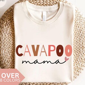 Puede incluir: Camiseta color crema con la frase "CAVAPOO mama" en letras marrones, rosas y negras, con un pequeño corazón rosa. La camiseta se muestra sobre una cesta tejida. También se ve el texto "OVER 8 COLORS".