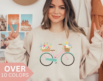 Op de afbeelding: Een licht beige sweatshirt met een roze fiets met een mand vol bloemen aan de voorkant. De tekst "OVER 10 COLORS" staat linksonder in de afbeelding.