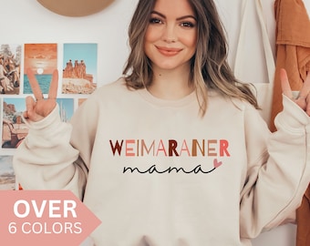 May include: A light beige sweatshirt with the text "WEIMARANER mama" in pink, brown, and orange letters with a small pink heart.