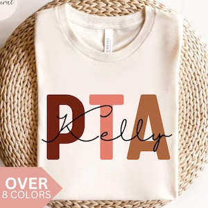 May include: A white t-shirt with the text "PTA Kelly" in brown, pink, and orange letters. The letters are stylized with a rounded font. The text is centered on the shirt.