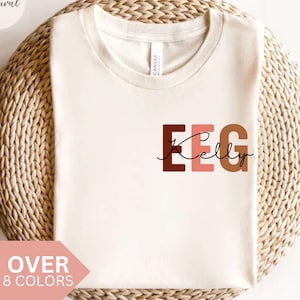 Può includere: Una maglietta bianca con il testo "EEG Kelly" in lettere marroni, rosa e beige. Le lettere sono stilizzate con un font arrotondato e il nome "Kelly" è scritto in corsivo.