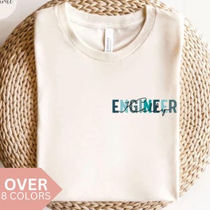 Op de afbeelding: Crèmekleurig T-shirt met het woord "ENGINEER" in teal en marineblauw, met een cursief "ingly" eronder. Het shirt wordt weergegeven op een geweven, cirkelvormig oppervlak. Tekst linksboven zegt "Natural" en linksonder "OVER 8 COLORS".